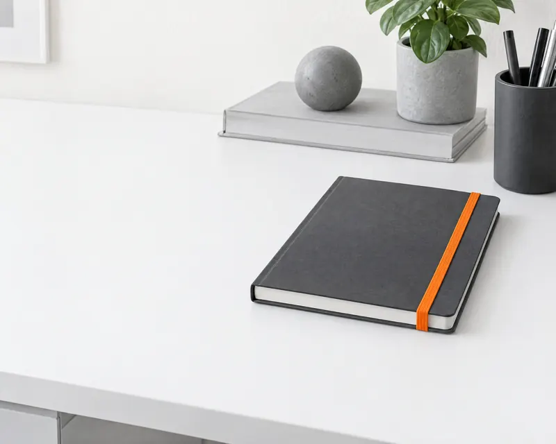 A closed graphite notebook on a white desk, held shut by an orange elastic band.